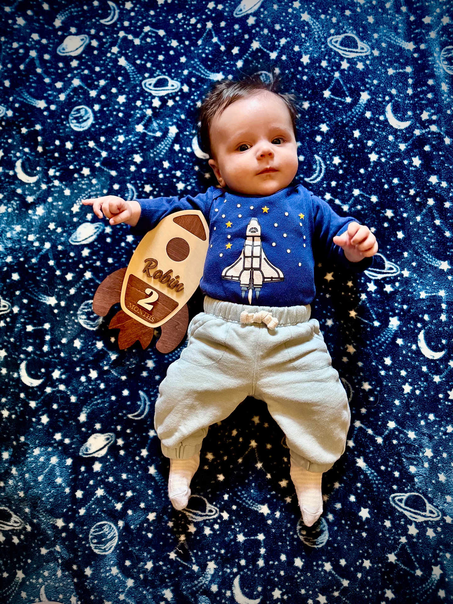 Robin is registered to the contest to win money with this photo: baby, infant, child, blanket, space_theme, rocket, wooden_sign, blue_shirt, pants, socks, lying_down, stars, planets, moon, cute, portrait, indoors, young_child, comfortable, adorable