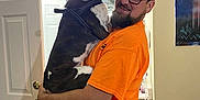 Nevaeh joined the competition — help win amazing prizes! man, dog, indoor, orange_shirt, glasses, beard, holding, door, painting, carpet, bed, light_fixture, mirror, white_door, black_and_white_dog, happy, casual, jeans, pet, home