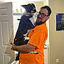 man, dog, indoor, orange_shirt, glasses, beard, holding, door, painting, carpet, bed, light_fixture, mirror, white_door, black_and_white_dog, happy, casual, jeans, pet, home