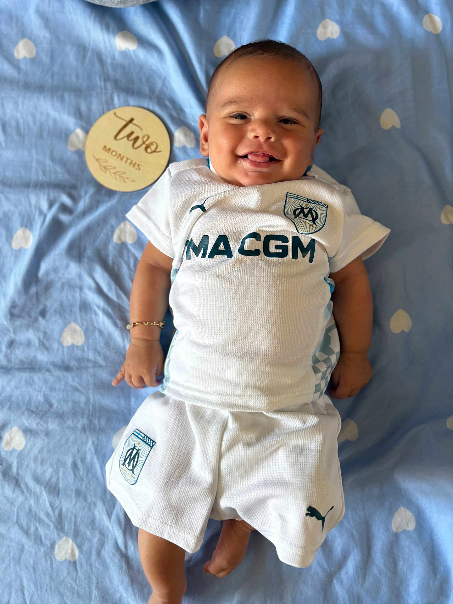 Tony a rejoint le concours — aidez-le/la à gagner de superbes lots ! baby, infant, smiling, white_clothing, sportswear, blue_blanket, heart_pattern, wooden_plaque, text, two_months, lying_down, happy, cute, infant_boy, bracelet, tongue_out, indoor, portrait, child, person