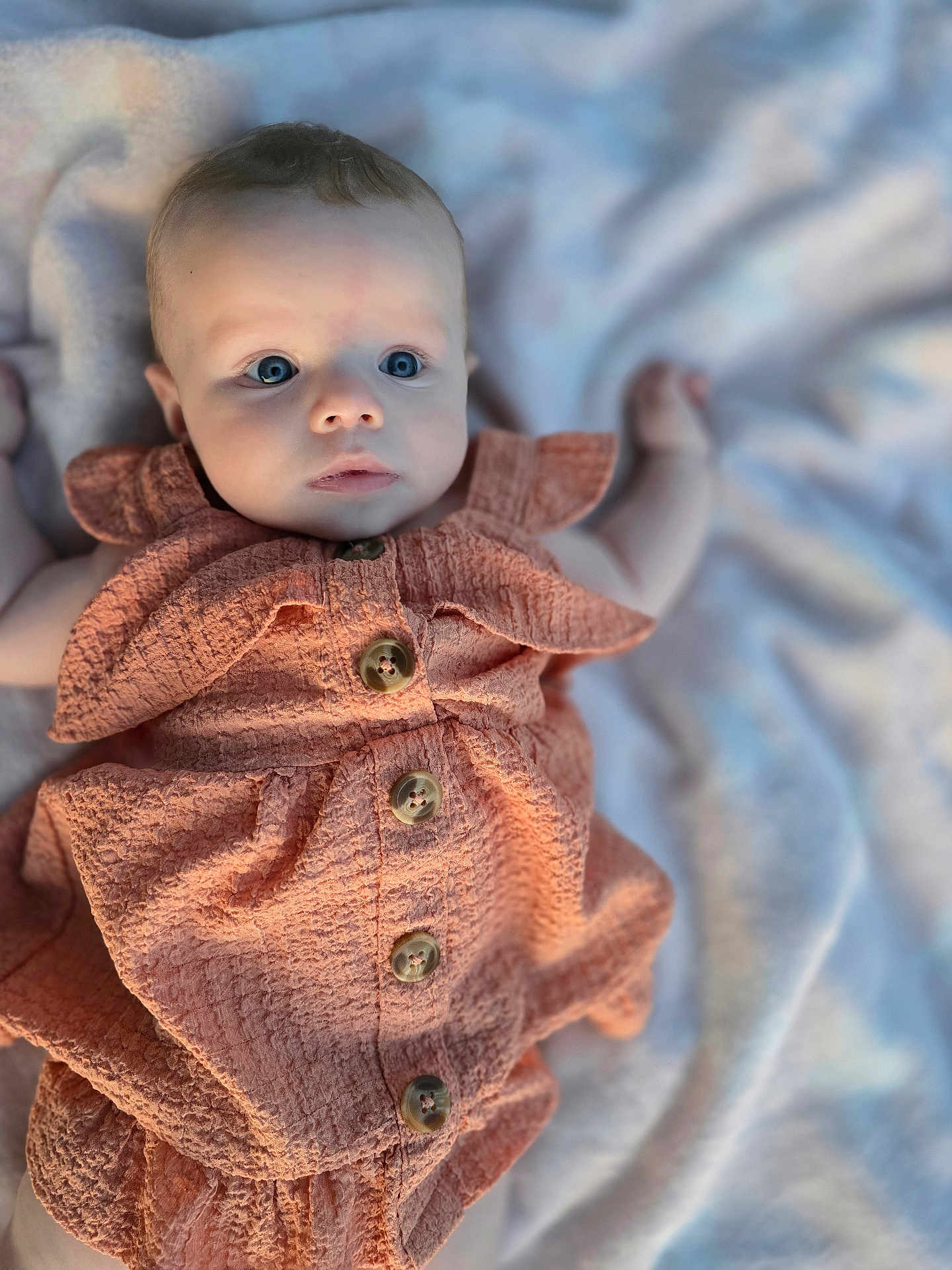 Brielle is registered to the contest to win money with this photo: baby, infant, child, blue_eyes, dress, buttoned_clothing, blanket, soft_texture, curious_expression, laying_down, closeup, portrait, cute, skin, face, small_hands, indoor, comfortable, young_child, apparel