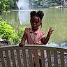 beads, bench, child, cute, daylight, expression, floral_pattern, girl, greenery, hair_buns, lake, nature, outdoor, pavement, peace_sign, playful, portrait, summer_clothing, trees, water_fountain