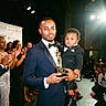 man, child, tuxedo, award, trophy, stage, applause, crowd, celebration, event, formal_wear, father, son, black_suit, smile, indoor, people, lighting, portrait, ceremony