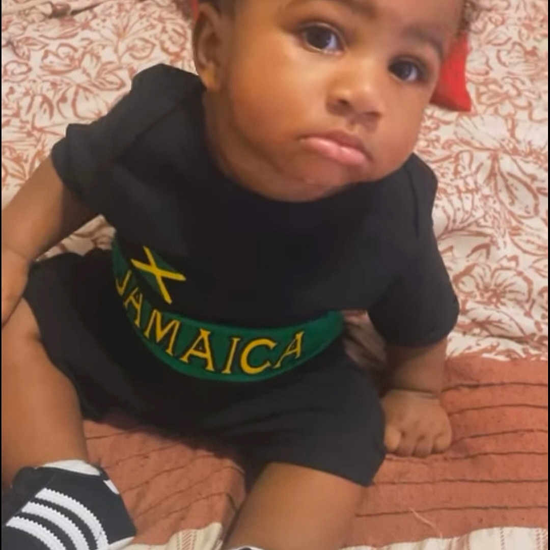 D'Montae is registered to the contest to win money with this photo: toddler, child, baby, indoor, bed, clothing, sneakers, black_shirt, jamaica, pillow, blanket, patterned, cute, face, expression, sitting, footwear, young_child, person, portrait