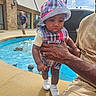 child, toddler, baby, plaid, hat, white_shoes, adult_hand, tattoo, fountain, water, outdoor, building, sidewalk, person, man, casual_clothing, daylight, expression, holding, closeup