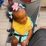 Nyonna Carswell joined the competition — help win amazing prizes! black_strap, casual, chair, child, curly_hair, floor, hair_bows, happy, indoor, joyful, legs, light_blue_clothing, playful, seated, shoes, smiling, stroller, toddler, wooden_furniture, yellow_bib