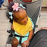 black_strap, casual, chair, child, curly_hair, floor, hair_bows, happy, indoor, joyful, legs, light_blue_clothing, playful, seated, shoes, smiling, stroller, toddler, wooden_furniture, yellow_bib