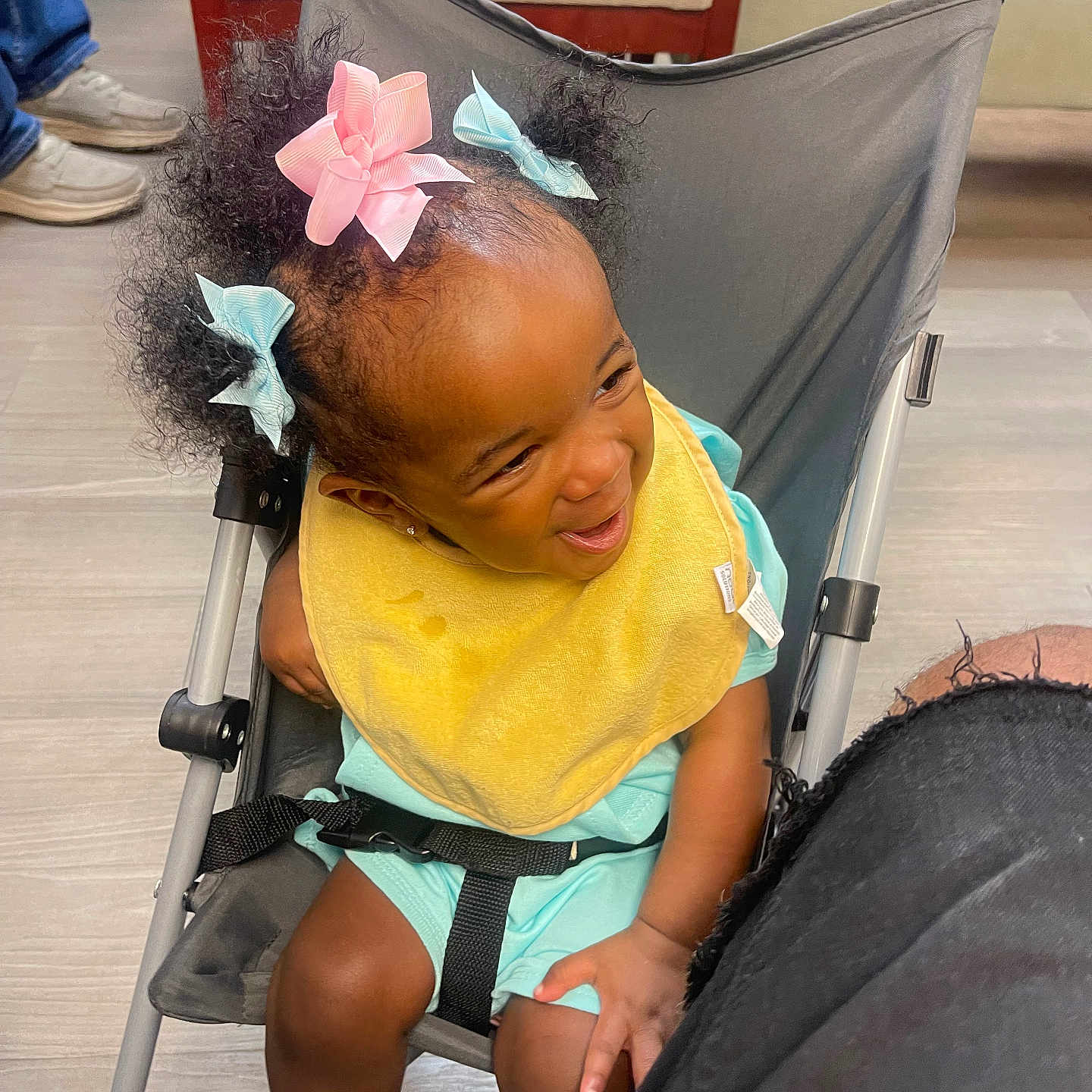 Nyonna Carswell joined the competition — help win amazing prizes! black_strap, casual, chair, child, curly_hair, floor, hair_bows, happy, indoor, joyful, legs, light_blue_clothing, playful, seated, shoes, smiling, stroller, toddler, wooden_furniture, yellow_bib