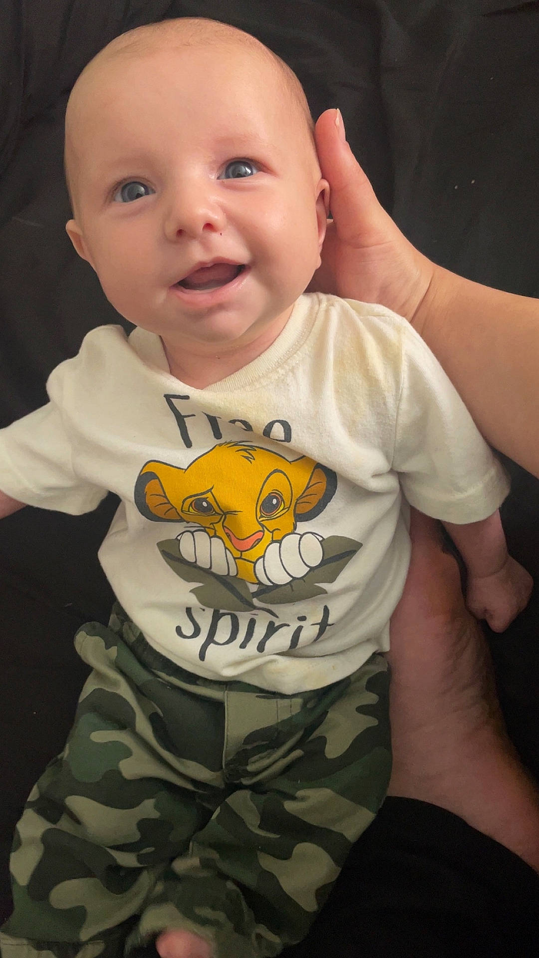 baby, blue_eyes, camouflage_pants, cartoon, child, clothing, cute, face, hand, happy, indoors, infant, lion, person, portrait, shirt, sitting, smiling, supporting_hand, toddler