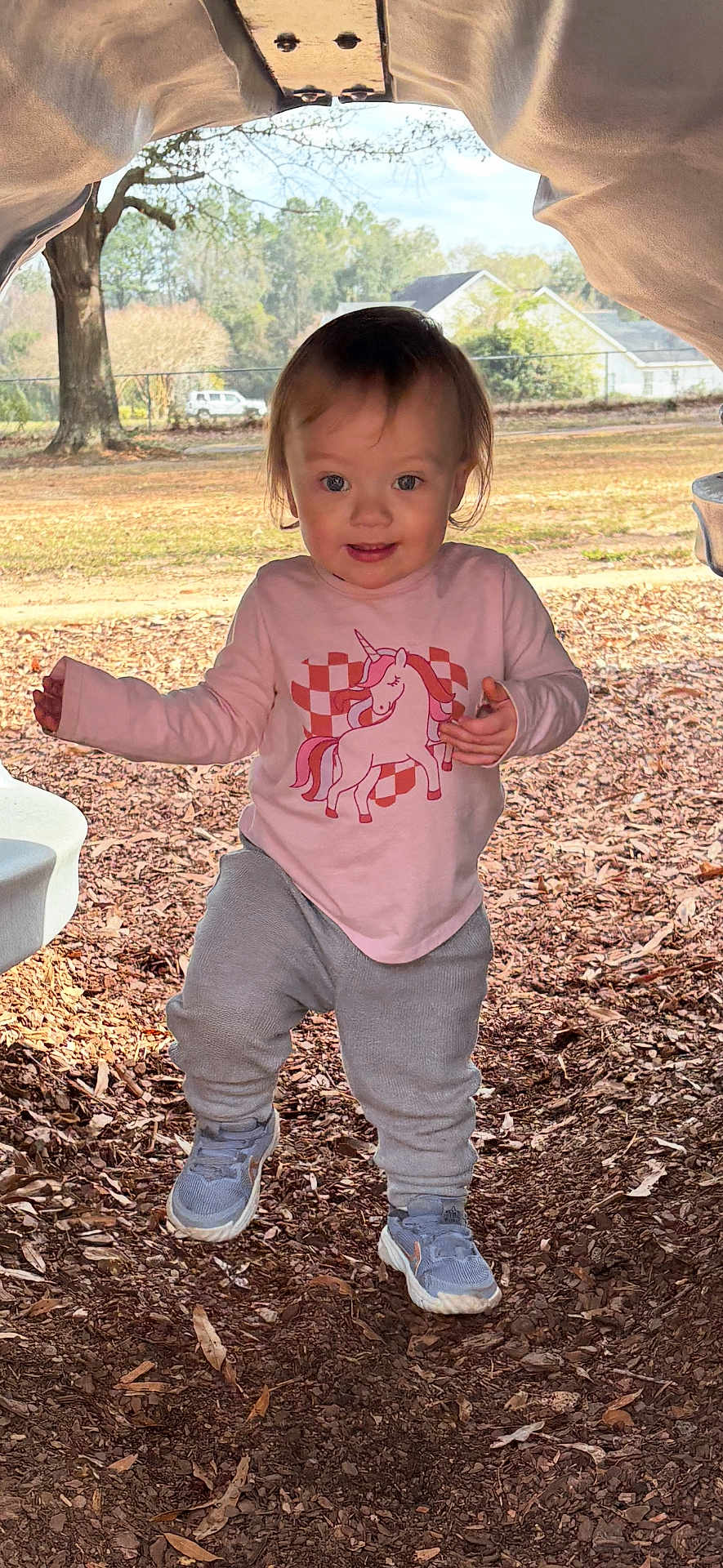 Jaylynn is registered to the contest to win money with this photo: toddler, child, outdoor, playground, unicorn_shirt, gray_pants, sneakers, leaves, wood_chips, smiling, face, person, daylight, tree, background, play_tunnel, nature, casual_clothing, happy, young_child