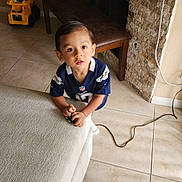 Milán joined the competition — help win amazing prizes! toddler, child, indoor, couch, toy, floor, tile_floor, bench, stone_wall, fireplace, sports_jersey, curious, looking_up, hands, white_socks, brown_hair, living_room, power_cord, yellow_toy, furniture