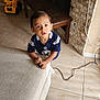 toddler, child, indoor, couch, toy, floor, tile_floor, bench, stone_wall, fireplace, sports_jersey, curious, looking_up, hands, white_socks, brown_hair, living_room, power_cord, yellow_toy, furniture