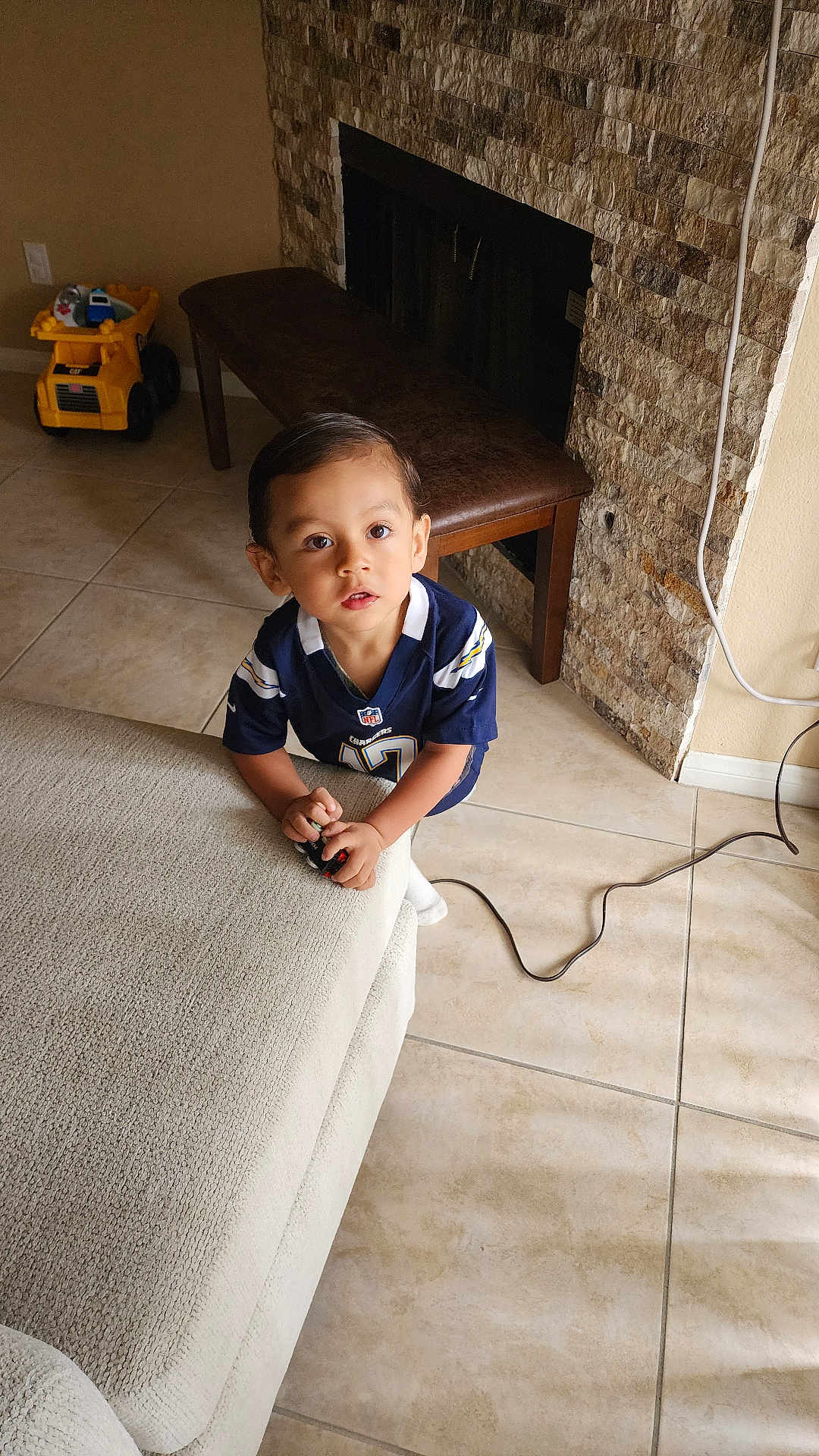 Milán joined the competition — help win amazing prizes! toddler, child, indoor, couch, toy, floor, tile_floor, bench, stone_wall, fireplace, sports_jersey, curious, looking_up, hands, white_socks, brown_hair, living_room, power_cord, yellow_toy, furniture