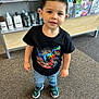 child, boy, tshirt, dinosaur, jeans, shoes, carpet, indoor, shelf, magazines, hair_products, black_tshirt, standing, cute, young_child, smiling, short_hair, clothing, retail, store