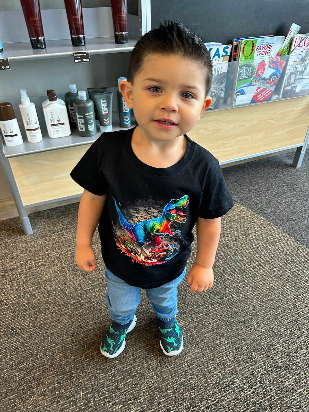 Isaiah is registered to the contest to win money with this photo: child, boy, tshirt, dinosaur, jeans, shoes, carpet, indoor, shelf, magazines, hair_products, black_tshirt, standing, cute, young_child, smiling, short_hair, clothing, retail, store