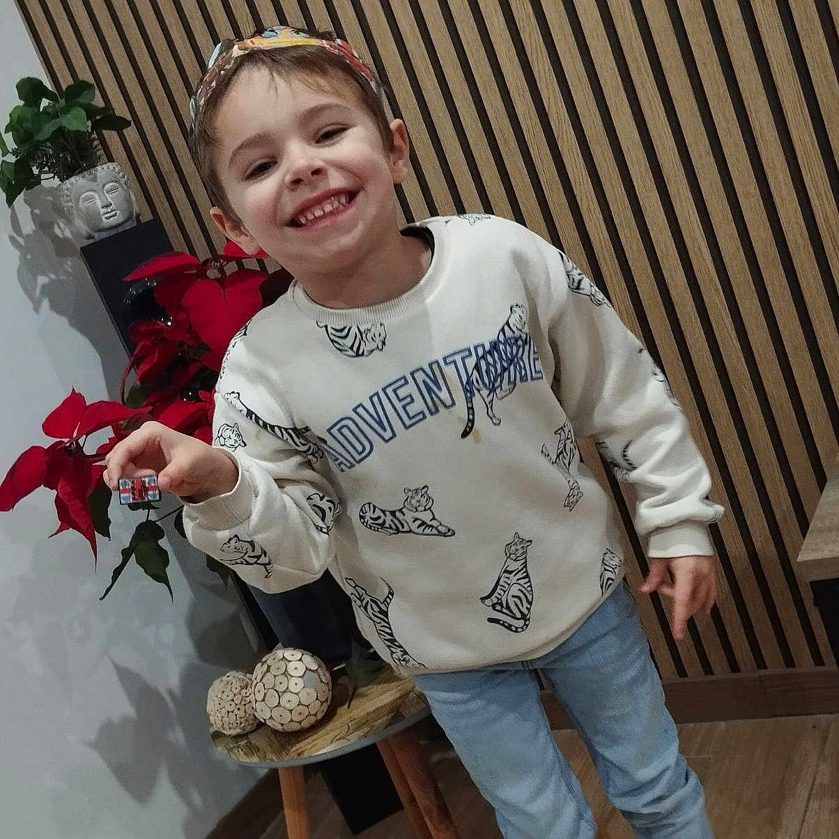 Leo a rejoint le concours — aidez-le/la à gagner de superbes lots ! casual_clothing, child, decor, floor, furniture, happy, headband, holding_object, home, indoor, jeans, person, plant, poinsettia, portrait, smiling, sweatshirt, table, tiger_pattern, wooden_wall
