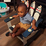 Kaleb is registered to the contest to win money with this photo: toddler, child, rocking_chair, indoor, wooden_chair, smiling, clothing, shorts, shirt, floor, toy, cabinet, books, furniture, decor, person, happy, baby, face, foot