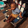 toddler, child, rocking_chair, indoor, wooden_chair, smiling, clothing, shorts, shirt, floor, toy, cabinet, books, furniture, decor, person, happy, baby, face, foot