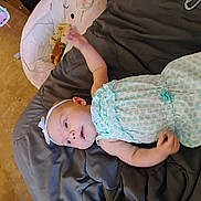 Natalie is registered to the contest to win money with this photo: baby, infant, headband, blue_outfit, lying_down, blanket, soft_toy, play_mat, indoor, child, arm_raised, curious, cute, floor, patterned_clothing, white_headband, baby_girl, soft_lighting, relaxed, cozy