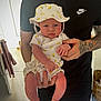 baby, infant, child, person, tattoo, hat, clothing, lemon_pattern, black_shirt, nike_logo, hand, indoor, carpet, basket, furniture, adult, holding, skin, expression, cute