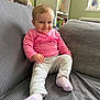 toddler, child, smiling, pink_top, light_pants, socks, couch, gray_couch, stuffed_animal, toys, bookshelf, books, indoors, home, playful, happy, person, furniture, window, daylight