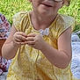 child, girl, glasses, yellow_dress, sitting, outdoor, grass, snack_bag, blanket, happy, smiling, curly_hair, person, summer, playful, cute, young, casual, daytime, relaxed