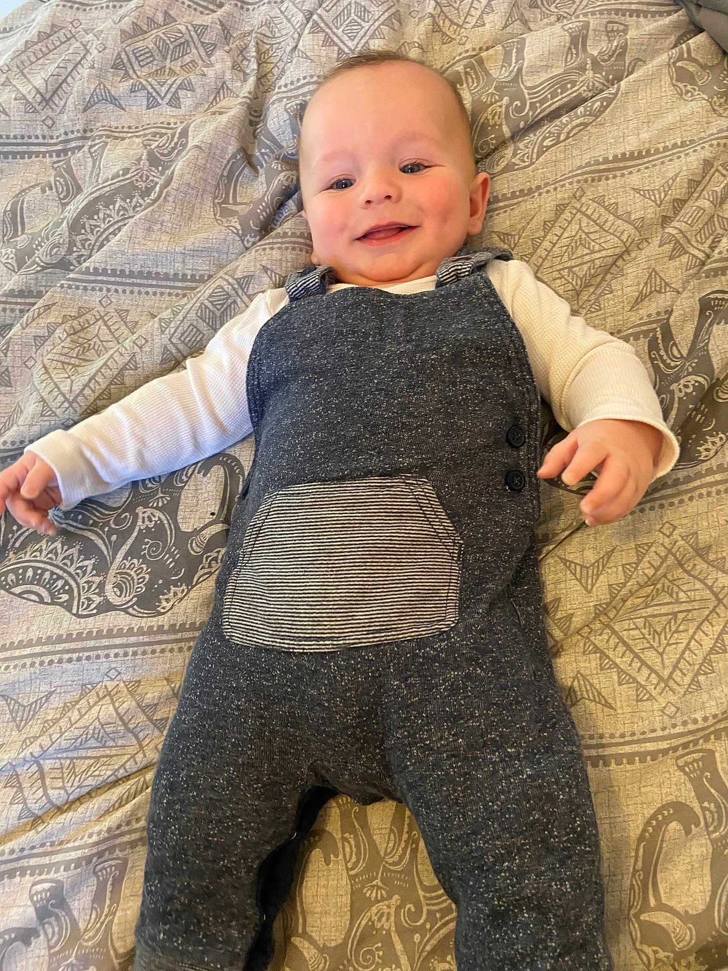 Arlo is registered to the contest to win money with this photo: baby, infant, smiling, overalls, clothing, bedspread, patterned, face, person, happy, cute, lying_down, child, indoor, portrait, skin, head, hand, textile, fabric