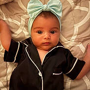 Alianna is registered to the contest to win money with this photo: baby, infant, headband, bow, pajamas, black_clothing, white_piping, bed, quilt, blanket, face, arms, wide_eyes, lying_down, indoors, soft_lighting, cute, child, person, portrait