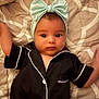 baby, infant, headband, bow, pajamas, black_clothing, white_piping, bed, quilt, blanket, face, arms, wide_eyes, lying_down, indoors, soft_lighting, cute, child, person, portrait