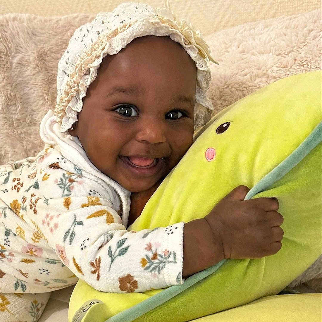 Nailah participe au concours pour gagner de l'argent avec cette photo : baby, bonnet, child, closeup, cozy, cuddling, cute, floral_pattern, green, happy, indoor, infant, lace, onesie, person, plush_pillow, portrait, smiling, soft_texture, toddler