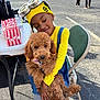 Pax is registered to the contest to win money with this photo: child, dog, puppy, yellow_clothing, hat, goggles, popcorn, table, chair, outdoor, smiling, happy, pavement, people, water_bottle, parking_lot, casual, playful, cute, holding