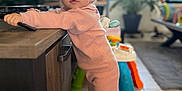Mya a rejoint le concours — aidez-le/la à gagner de superbes lots ! toddler, child, pink_pajamas, standing, indoor, wooden_counter, toy, plant, living_room, shelving, floor_tile, curious, person, baby, home_decor, furniture, footwear, natural_light, face, portrait