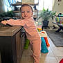 toddler, child, pink_pajamas, standing, indoor, wooden_counter, toy, plant, living_room, shelving, floor_tile, curious, person, baby, home_decor, furniture, footwear, natural_light, face, portrait