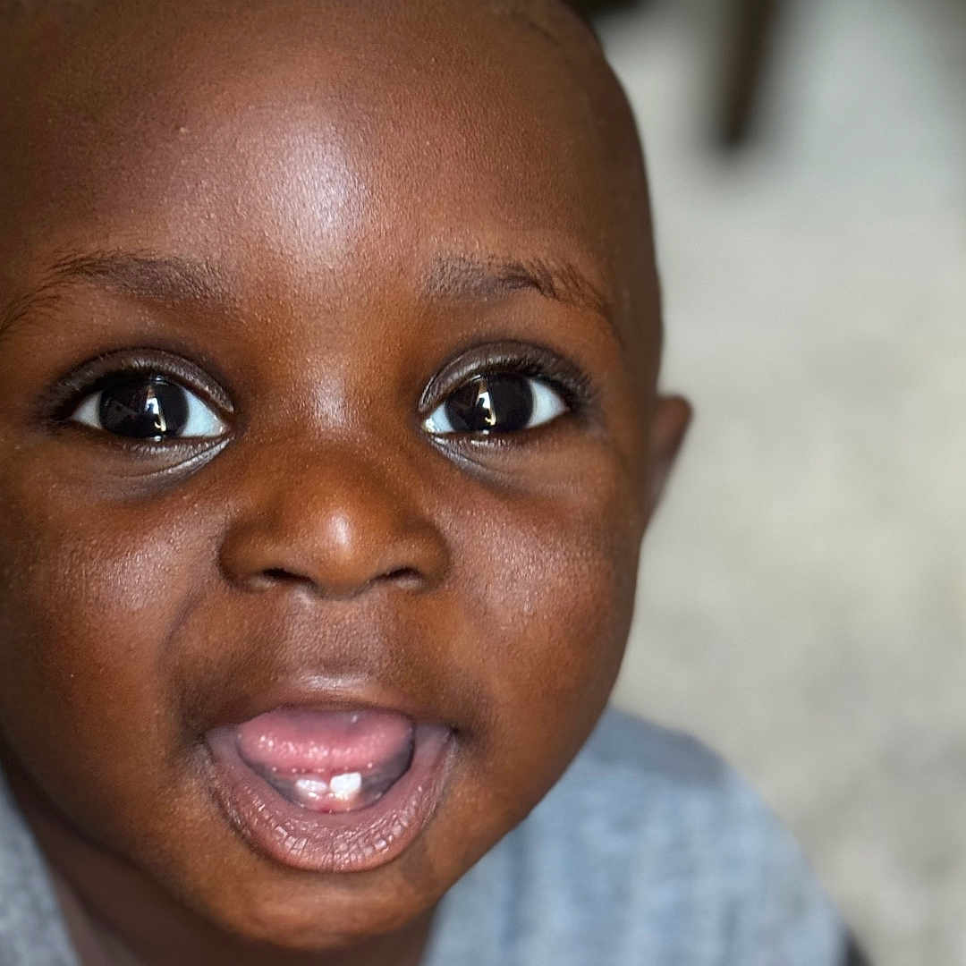 Muhammad is registered to the contest to win money with this photo: child, toddler, face, smiling, mouth_open, teeth, eyes, skin, portrait, indoor, clothing, gray_shirt, cute, happy, baby, person, head, young, expression, closeup