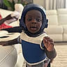 toddler, child, helmet, protective_vest, indoor, living_room, couch, carpet, furniture, curious, reaching, person, clothing, short_sleeves, plaid_shirt, black_skin, young_child, home, blurred_background, soft_lighting