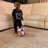 Muhammad is registered to the contest to win money with this photo: toddler, child, soccer_ball, rug, indoor, living_room, couch, plant, black_clothing, graphic_tshirt, footwear, portrait, carpet, home_decor, serious_expression, young_child, furniture, person, standing, toy