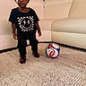 child, soccer_ball, carpet, black_clothing, indoor, sofa, plant, footwear, person, young_child, sport, toy, room, home, standing, cute, smiling, fashion, casual, living_room
