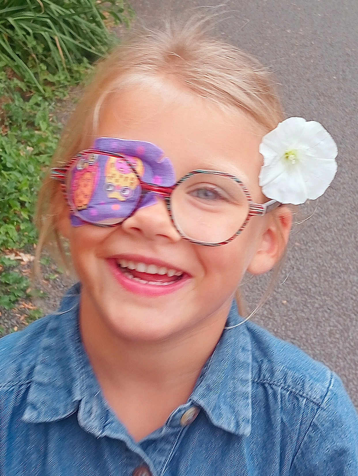 Leana participe au concours pour gagner de l'argent avec cette photo : accessory, blonde_hair, casual_clothing, child, denim_shirt, eye_patch, eyewear, face, flower, glasses, greenery, happy, head, nature, outdoor, pathway, person, portrait, smile, young