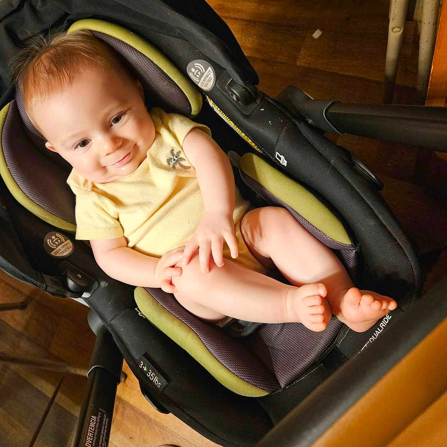Brantley is registered to the contest to win money with this photo: baby, child, infant, smiling, portrait, car_seat, stroller, seated, indoors, wooden_floor, yellow_shirt, barefoot, toes, happy, looking_at_camera, close_up, baby_gear, warm_lighting, home, person