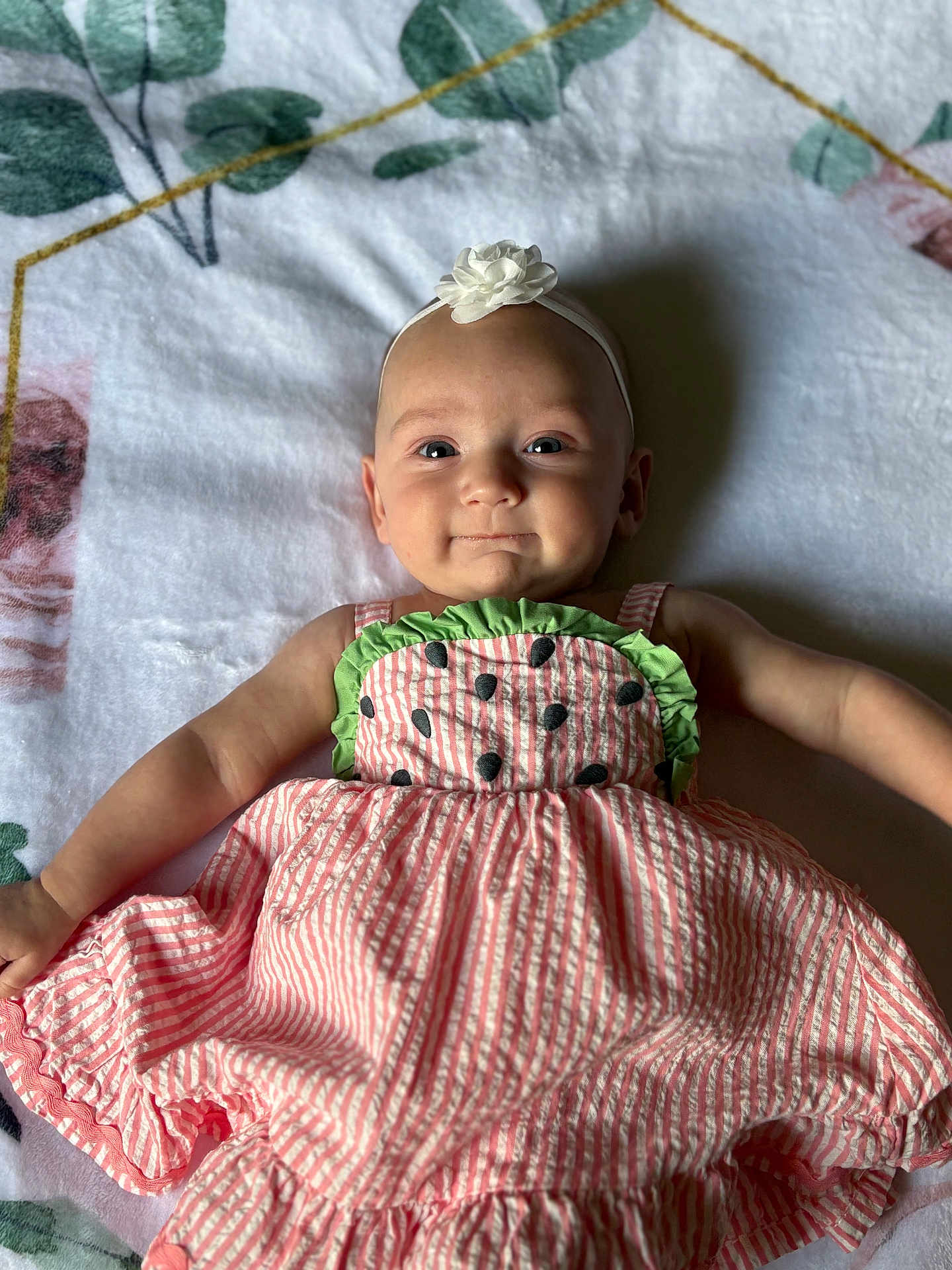 Addison is registered to the contest to win money with this photo: baby, infant, child, smiling, dress, headband, flower, blanket, patterned_clothing, pink, green, laying_down, cute, happy, indoor, portrait, face, skin, young, person