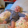 baby, smiling, infant, pillow, floral_pattern, bedroom, blanket, happy, face, person, clothing, sitting, head, cute, interior, furniture, relaxing, child, tongue_out, cozy
