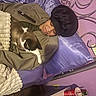 bed, blanket, bonnet, cat, clothing, cup, decor, furniture, headboard, indoor, jacket, person, pillow, purple, relaxed, remote_control, resting, sleeping, table, water_bottle