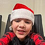 beige_wall, black_chair, casual_clothing, child, close_up, earrings, face, festive, freckles, happy, holiday, indoor, person, plaid_shirt, portrait, red_and_black, red_hat, santa_hat, smiling, young