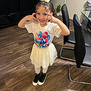 Bianca is registered to the contest to win money with this photo: child, girl, smiling, happy, birthday, tshirt, skirt, white, black_shoes, curly_hair, bow, indoor, wooden_floor, chair, sofa, plant, home, playful, pose, person