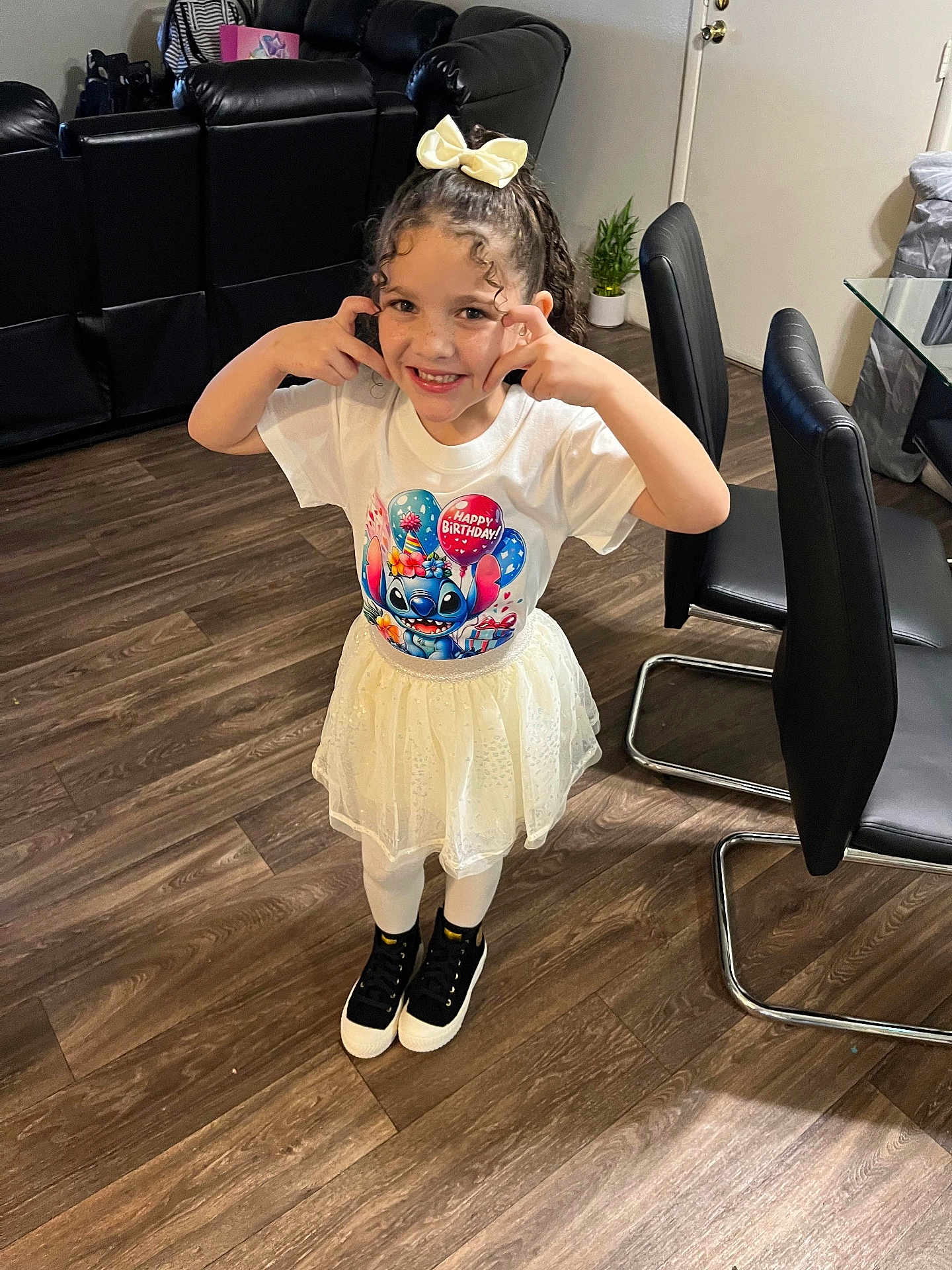 Bianca is registered to the contest to win money with this photo: child, girl, smiling, happy, birthday, tshirt, skirt, white, black_shoes, curly_hair, bow, indoor, wooden_floor, chair, sofa, plant, home, playful, pose, person