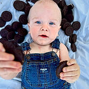 Jack is registered to the contest to win money with this photo: baby, child, blue_eyes, denim_overalls, cookies, oreo, snack, food, hand, face, infant, cute, lying_down, blue_background, skin, expression, portrait, close_up, holding, playful