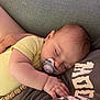 adult_hand, baby, close_up, clothing, comfort, face, hand, human, indoor, nap, pacifier, peaceful, person, resting, shirt, skin, sleeping, soft_lighting, text_on_clothing, yellow_onesie