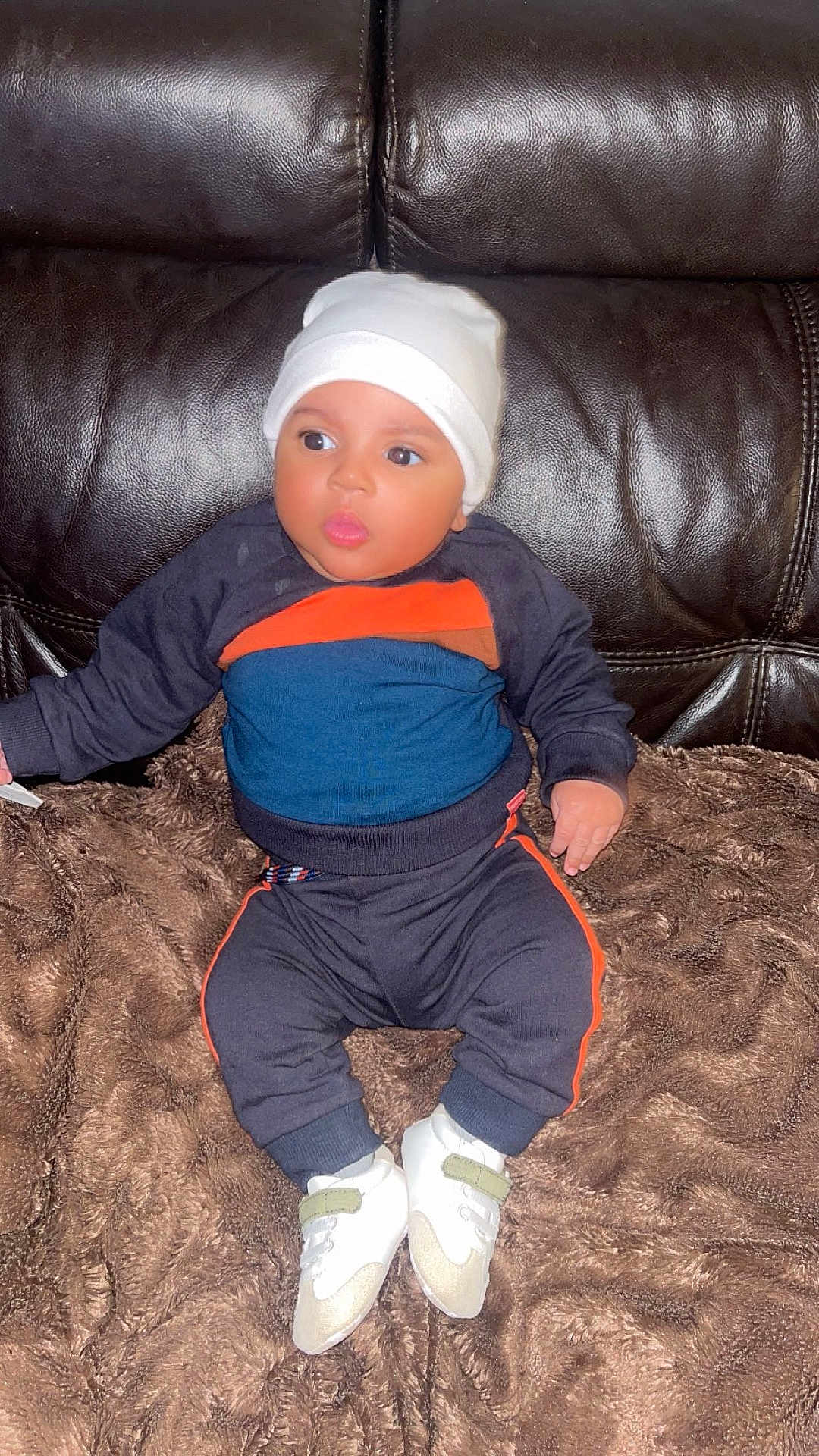 Loann a rejoint le concours — aidez-le/la à gagner de superbes lots ! baby, child, infant, sitting, blanket, leather_couch, white_hat, white_shoes, sportswear, curious, indoor, cozy, cute, small_hands, wide_eyes, puckered_lips, dark_background, soft_texture, comfortable, portrait