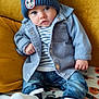 baby, child, blue_hat, mickey_mouse_patch, denim_jacket, striped_shirt, jeans, white_shoes, blanket, pumpkin_pattern, black_cat_pattern, couch, yellow_couch, seated, indoors, cute, portrait, cozy, fall_theme, casual_clothing