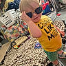 Braxley is registered to the contest to win money with this photo: blanket, carpet, casual, child, clothing, face, floor, hand, home, indoor, laundry_basket, person, plush_toy, quilt, sneakers, standing, sunglasses, toddler, toy, yellow_shirt